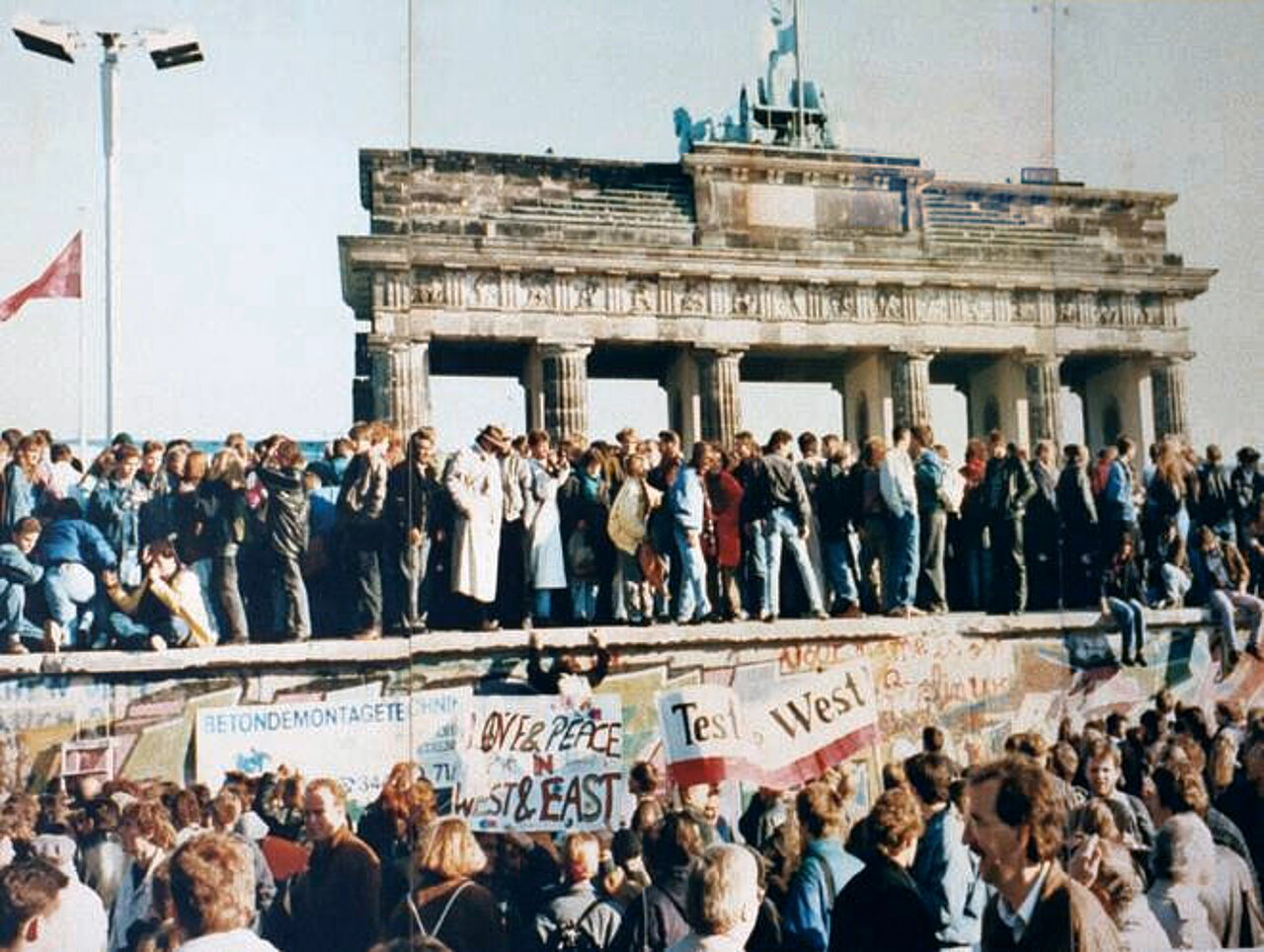 Farbfoto: Eine große Gruppe an Menschen steht vor und auf der Berliner Mauer, im Hintergrund ist das Brandenburger Tor zu sehen.