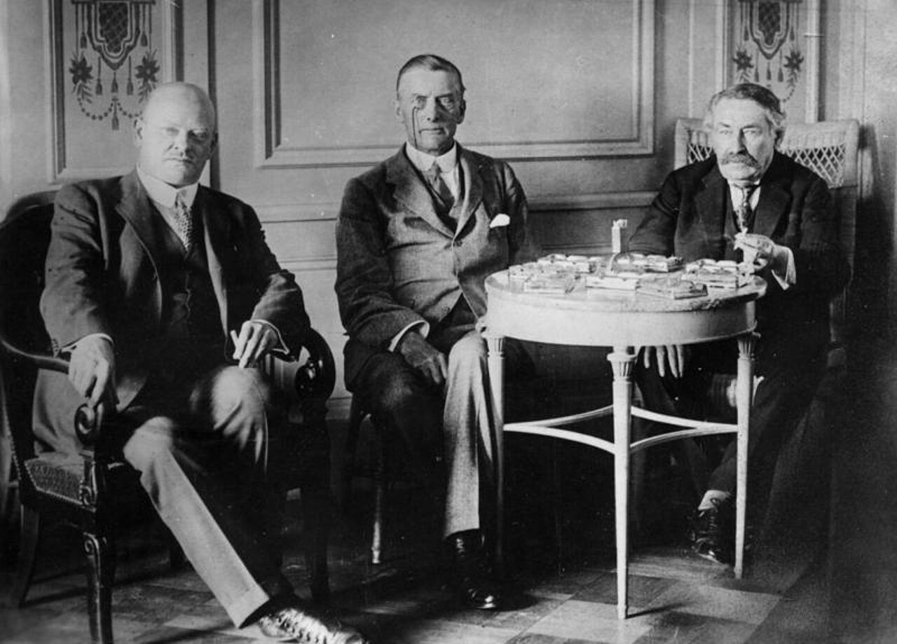Schwarz-weiß-Foto: Drei elegant gekleidete Männer sitzen nebeneinander.