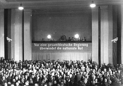 Schwarz-weiß-Foto: Menschengruppe in historischem Saal, auf einer Empore weitere Menschen an der ein Banner mit der Aufschrift: "Nur eine gesamtdeutsche Regierung überwindet die nationale Not" hängt.