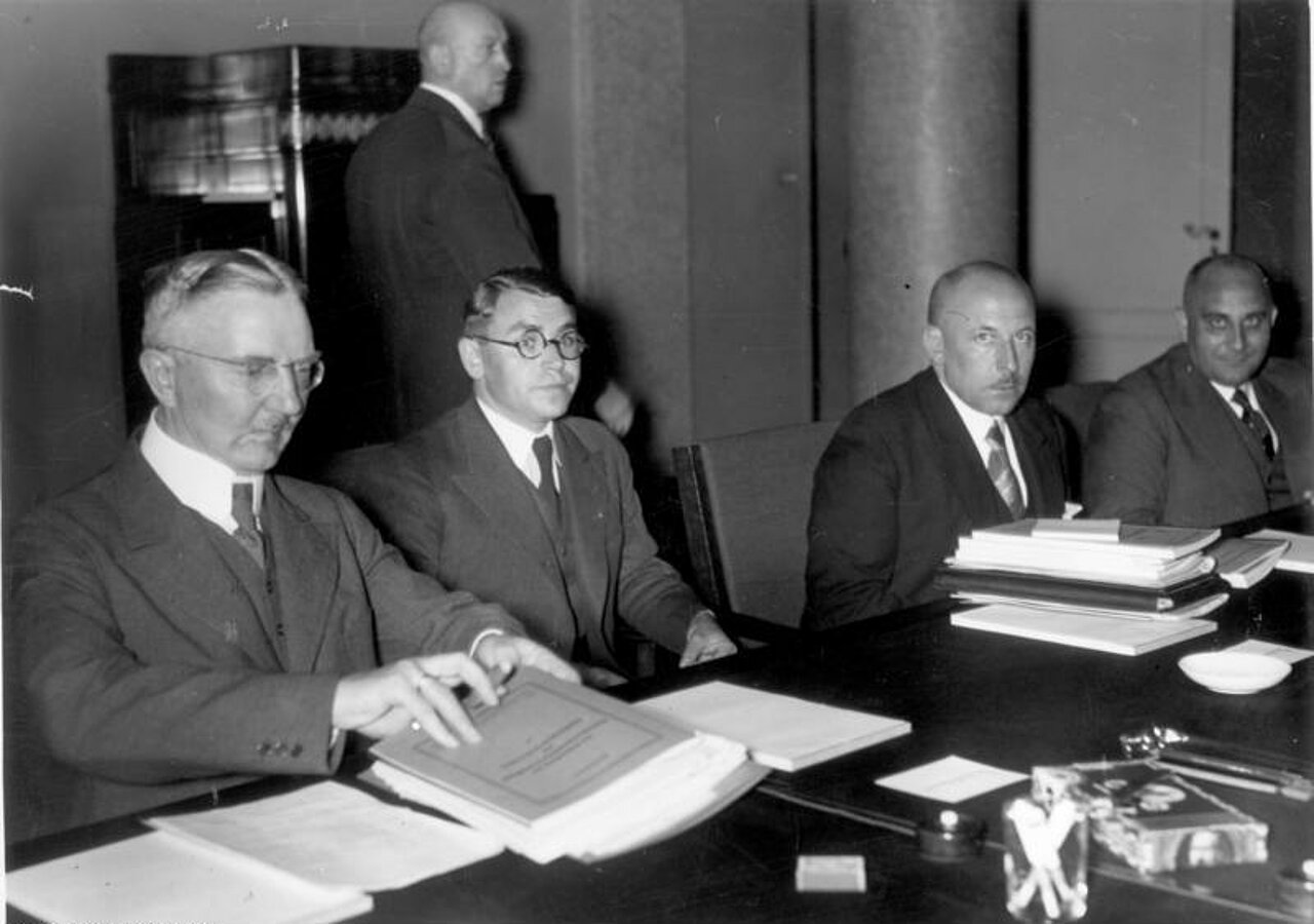 Schwarz-weiß-Foto: Vier Herren in Anzügen sitzen am Tisch vor Schriftstücken. Ein fünfter Mann steht hinter ihnen.