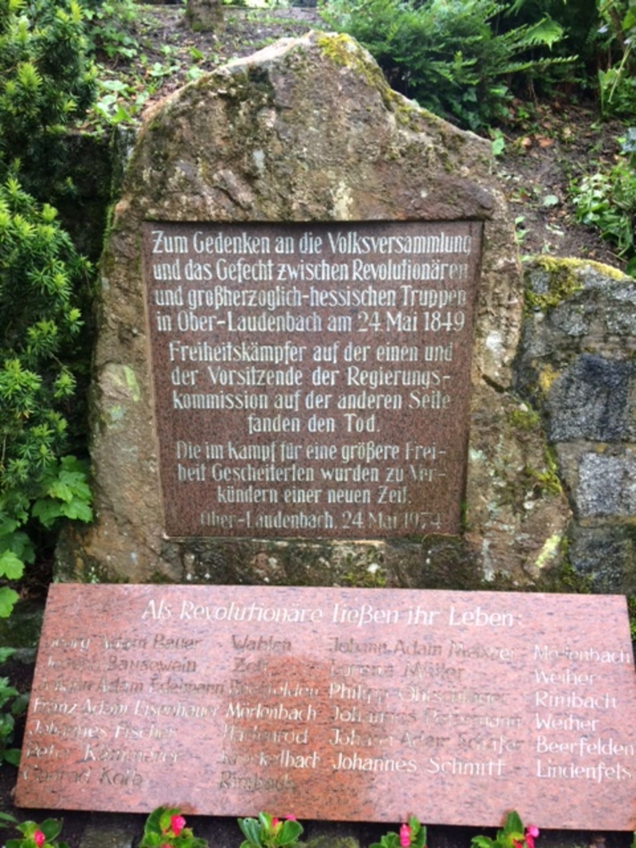 Gedenkstein mit Namenstafel vor grüner Kulisse.