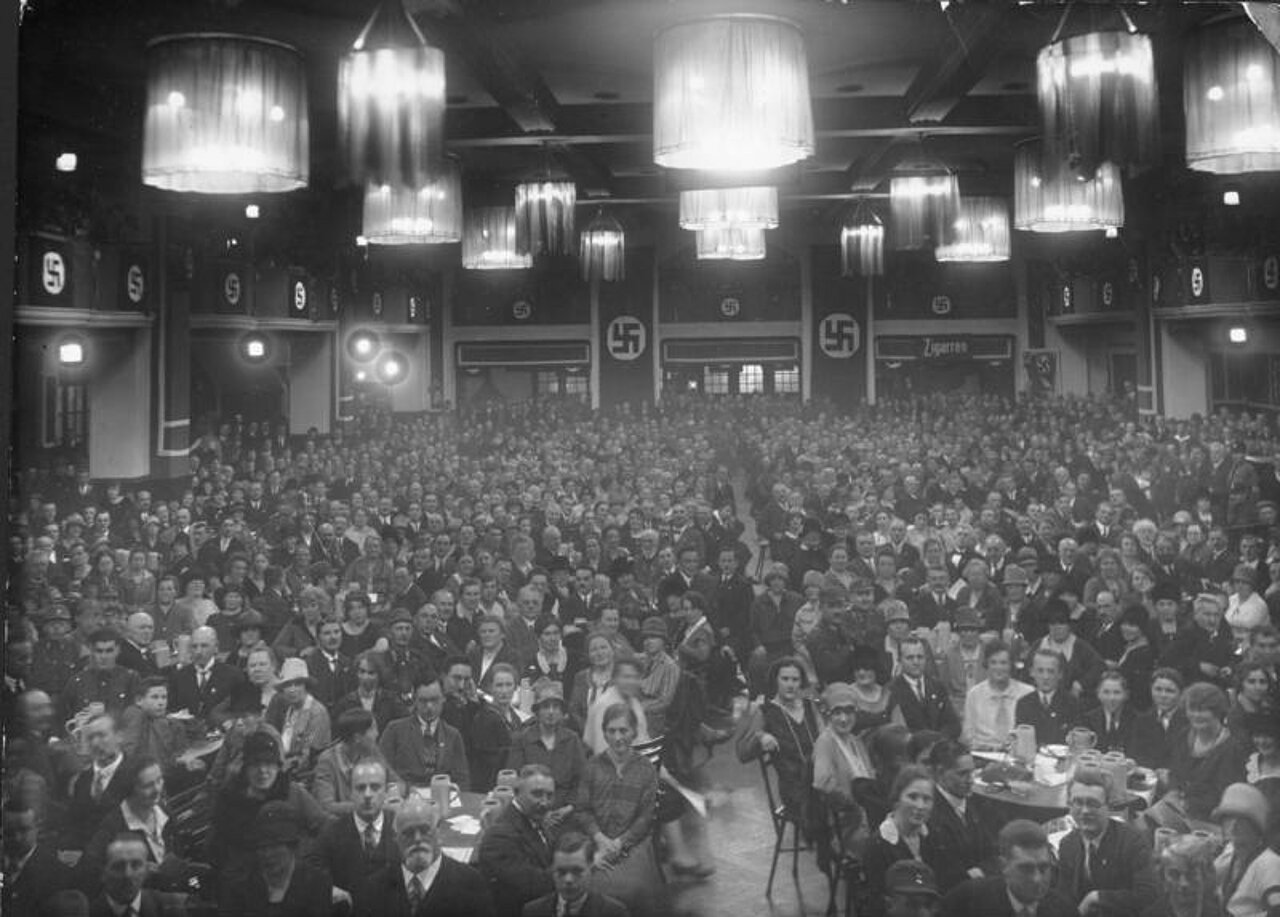 Schwarz-weiß-Foto: Viele Menschen sitzen an Tischen in einem Raum mit Kronleuchtern an der Decke und Hakenkreuz-Fahnen.